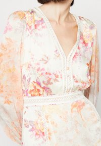 Robe longue à manches longues en tissu transparent, avec un décolleté en V, des détails de boutons et des accents en dentelle dans des teintes de rose, pêche et crème.