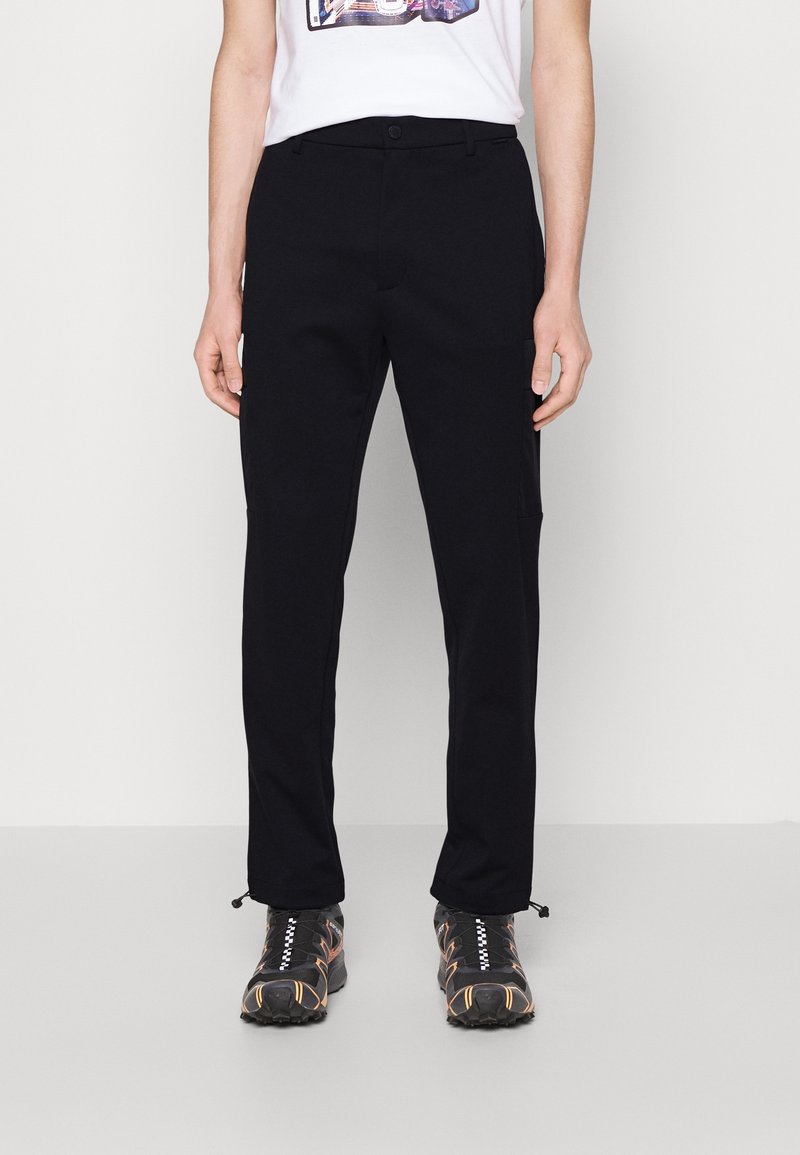 Personne portant un pantalon noir droit avec des cordons au bas et des chaussures de sport noires avec des détails orange.