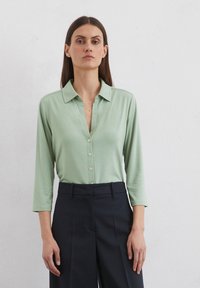 Light green, long-sleeve collared shirt with a button-down front, made from smooth fabric. Paired with dark, tailored trousers.