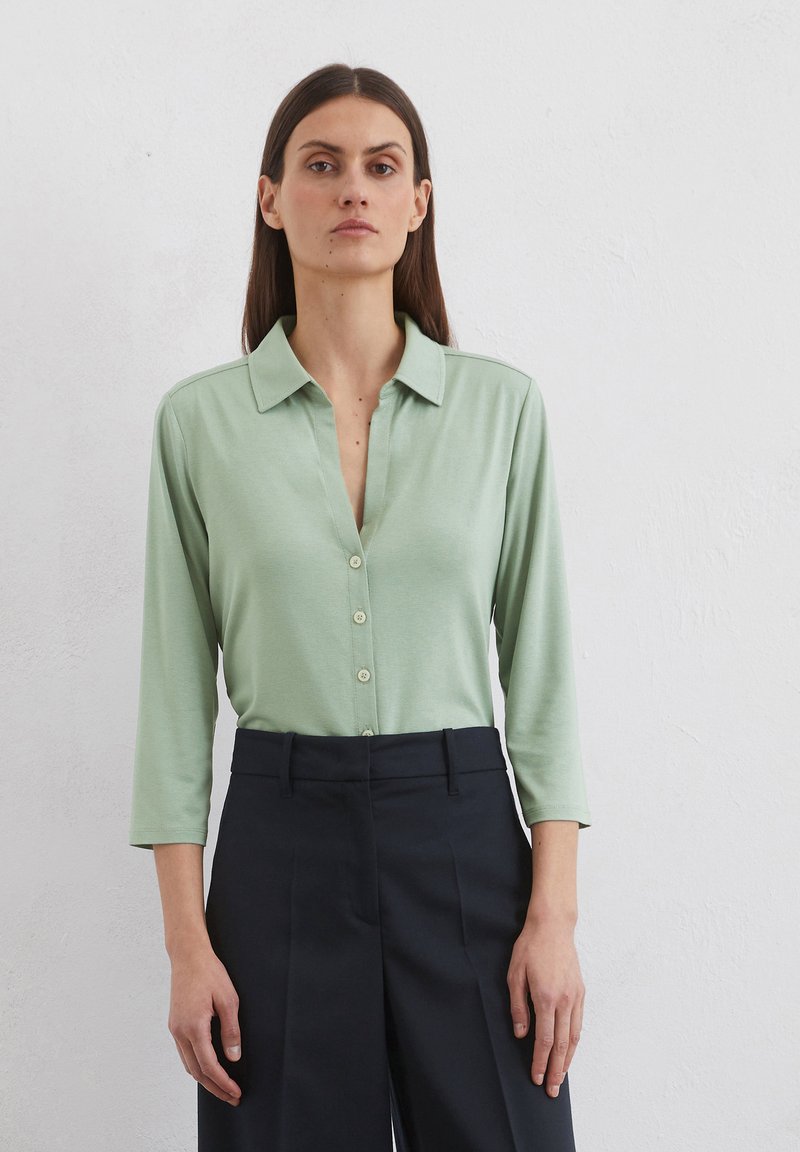 Light green, long-sleeve collared shirt with a button-down front, made from smooth fabric. Paired with dark, tailored trousers.
