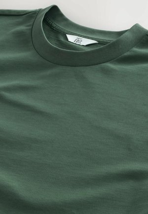 Camiseta de algodón verde oscuro con cuello redondo, textura suave y una etiqueta que muestra "NEXT EDIT" cerca del cuello.