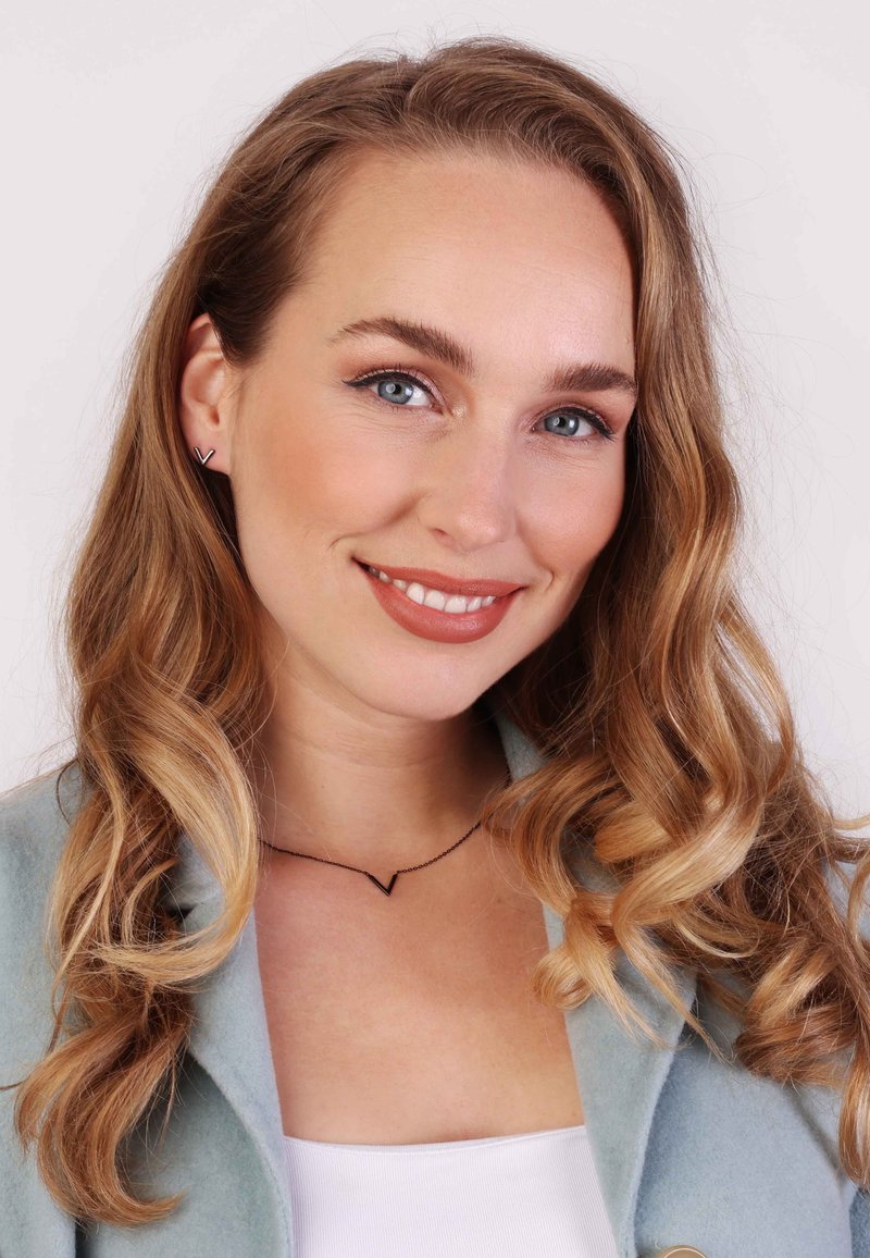 Schwarze V-förmige Halskette an einer Frau mit welligem, blonden Haar, die eine hellblaue Jacke und ein weißes Oberteil trägt, lächelnd vor einem neutralen Hintergrund.