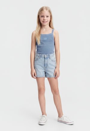 Νεαρό κορίτσι στέκεται, φοράει μπλε αμάνικη μπλούζα με το γράμμα "GAP", ανοιχτόχρωμα τζιν σορτς και άσπρα αθλητικά παπούτσια σε απλό λευκό φόντο.