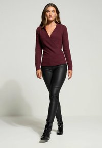 Frau, die in einem burgunderfarbenen gestrickten Wickelsweater, schwarzen Lederhosen und schwarzen Knöchelschuhen vor einem schlichten hellen Hintergrund spaziert.