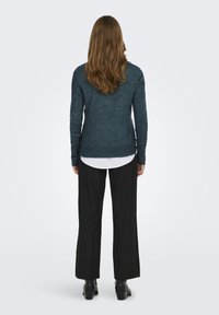 Dunkeltürkiser Pullover aus strukturiertem Stoff, mit taillierten Ärmeln und geripptem Saum, kombiniert mit weiten schwarzen Hosen und Stiefeletten. Ansicht von hinten.