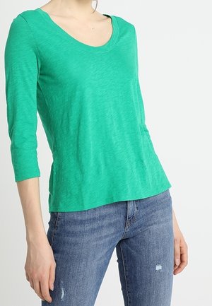 Frau trägt ein leuchtend grünes Shirt mit rundem Ausschnitt und dreiviertellangen Ärmeln sowie blaue, abgenutzte Jeans und steht vor einem schlichten hellen Hintergrund.