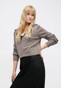 Rövidített barna puha kötött kapucnis pulóver cipzárral, bordázott mandzsettával és szegéllyel, kapucnival. Fekete szoknyával párosítva.