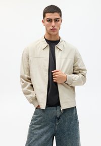 Lichtbeige bomberjack met een kraag, ribbelmanchetten en ritssluiting, gedragen over een donker shirt en denimshorts.