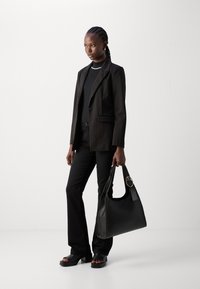 Zwarte blazer, aansluitend zwart topje en uitlopende zwarte broek. Houdt een grote zwarte handtas met een ronde gouden acccent vast. Glad, strak textuur overal.