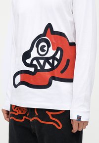 Λευκό πουκάμισο με μακριά μανίκια, με μεγάλο κόκκινο και μαύρο καρτουνίστικο σχέδιο γάτας, σε συνδυασμό με μαύρο παντελόνι που έχει αντίστοιχο σχέδιο περιγράμματος κόκκινης γάτας.