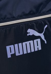 Gros plan sur un sac en tissu bleu marine avec une fermeture éclair argentée et le logo blanc Puma représentant un puma sautant et un texte en majuscules audacieuses.