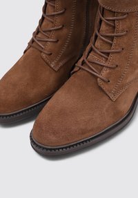 Botas de ante marrón con cordones, punta redondeada, suela de goma oscura contrastante y costuras detalladas en la parte superior.