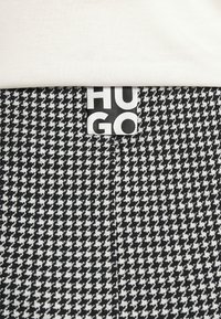 Tela de espiga en blanco y negro con una etiqueta blanca que presenta el texto "HUGO" en negrita y negro, parcialmente visible debajo de una prenda blanca.