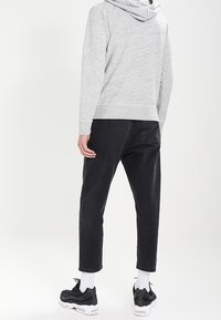 Person med ljusgrå huvtröja, svarta cropped byxor, vita strumpor och svarta sneakers som står mot en vit bakgrund, med ryggen mot kameran.