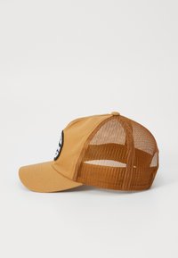 Beige cap med buet skygge, nettingpaneler på sidene og baksiden, og en rund svart patch med hvite grafikker foran.