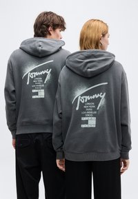 Δύο άτομα φορούν σκούρες γκρι μπλούζες με κουκούλα, με λευκό λογότυπο "Tommy" και ονόματα πόλεων τυπωμένα στην πλάτη, γυρισμένα προς την άλλη πλευρά μπροστά σε απλό φόντο.