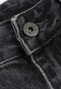 Czarny denim zbliżenie na metalowy guzik z grawerunkiem. Tkanina jest lekko zużyta, z widocznymi detalami szwów.