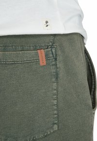 Groene broek van gestructureerde stof met een bruine leren label op de achterzak. Een wit shirt met een klein logo-label is gedeeltelijk zichtbaar erboven.