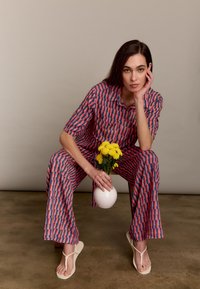 Een gestreepte kortarmige shirt en palazzo broek in roze en blauw, gecombineerd met witte teenslippers. Houdt een witte vaas met gele bloemen vast.