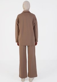 Bruin tweedelig outfit bestaande uit een lange mouwen top en een wijdvallende broek. Gemaakt van zachte stof, met een ontspannen pasvorm en een neutraal kleurenpalet.