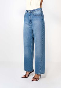 Široké modré džínsy vyrobené z denimového materiálu, s vysokým pásom, rovný strih a dvoma prednými vreckami. Skombinované s čiernymi sandálmi na vysokom podpätku.