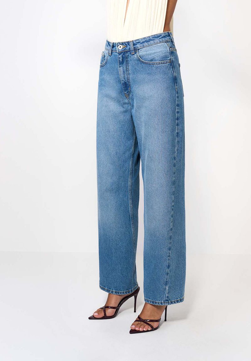 Široké modré džínsy vyrobené z denimového materiálu, s vysokým pásom, rovný strih a dvoma prednými vreckami. Skombinované s čiernymi sandálmi na vysokom podpätku.
