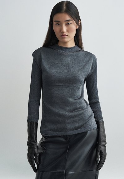 Maglione grigio a maniche lunghe con una texture aderente e colletto alto, abbinato a guanti in pelle nera e una gonna in pelle coordinata, su uno sfondo neutro.