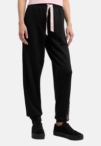 Schwarze Jogginghose mit schmalem Schnitt, elastischem Bund und pinkem Kordelzug. Sie bietet Seitentaschen und weißen Drucktext entlang des Beins.