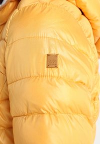 Manche d'une veste matelassée jaune vif avec un petit patch en cuir carré de la marque cousu sur le bras extérieur.