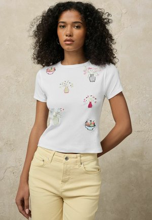 Kobieta o kręconych włosach w białej koszulce z kolorowymi, haftowanymi wazonami i kwiatami, w połączeniu z jasnżółtymi spodniami.