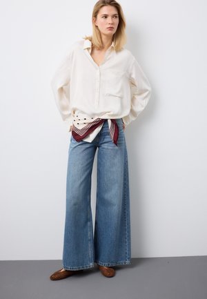 Mujer de pie con las manos detrás de la espalda, vistiendo una blusa blanca suelta, jeans de pierna ancha azules, bufanda burdeos con estampado blanco y zapatos marrones tejidos.