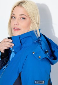 Sininen ulkotakki, jossa on korkea kaulus, kiristysnyörillinen huppu ja kiiltävä kangaspinta. Sisältää metalliset nepparit ja hihassa olevan logoplaatin.