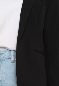 Černý blazer se strukturovaným designem, s jednou kapsou na přední straně, nošený přes bílé tričko, spárovaný se světle modrými denimovými džíny.