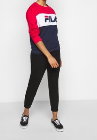 Persoon draagt een rood-wit-blauw FILA-sweatshirt, zwarte joggerbroek en zwarte sneakers, staand tegen een effen witte achtergrond.