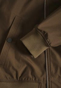 Brown bomber jacket with a smooth fabric finish, featuring a zipped front, ribbed cuffs, and a snap-button detail on the pocket.