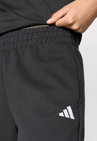 Μαύρο αθλητικό παντελόνι με ελαστική μέση, λεία υφή και λευκό λογότυπο στη αριστερή πλευρά. Κοντινή λήψη στην περιοχή της μέσης και χέρι που κρατάει το ύφασμα.