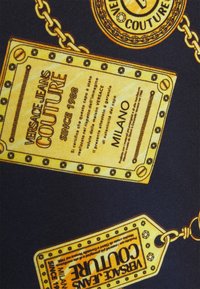 Tkanina Versace Jeans Couture w granatowym kolorze z wystającymi złotymi metkami z tekstem oraz ozdobnymi łańcuchami, prezentująca markowe detale i wzory.