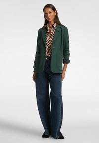 Donna che indossa un blazer verde, una camicia a fantasia geometrica, jeans larghi di colore blu scuro e scarpe nere, in piedi su uno sfondo bianco.