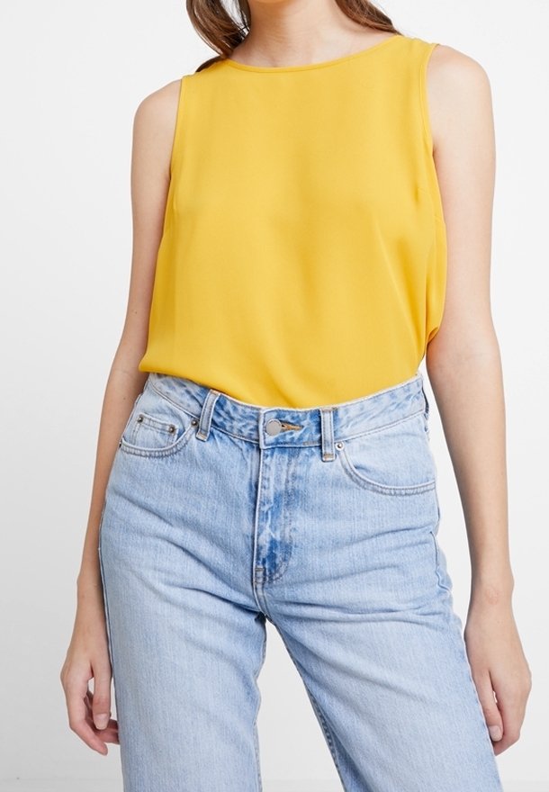 Żółta bezrękawnikowa bluzka z lekkiego materiału, połączona z jasnoniebieskimi dżinsami z wysokim stanem, które mają zapięcie na guzik i kieszenie z przodu.