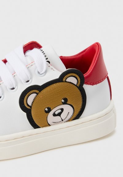 Vit sneaker med röd hälldetalj, med en texturerad björnhuvudsydd patch på sidan och vita snören. Gummisula och slät yta.