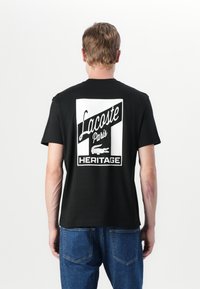 Svart bomulls t-skjorte med stort hvitt grafikk på baksiden, som viser "Lacoste Paris" og "HERITAGE" med et krokodilleikon.