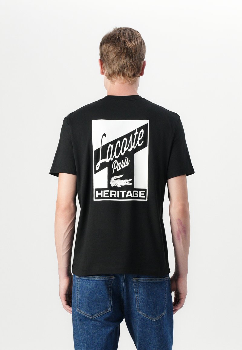 Svart bomulls t-skjorte med stort hvitt grafikk på baksiden, som viser "Lacoste Paris" og "HERITAGE" med et krokodilleikon.