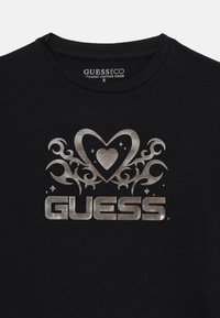Black cotton t-shirt featuring a metallic silver logo with heart and swirling designs, labeled "GUESS" beneath decorative elements.