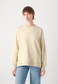 Felpa oversize di colore beige chiaro realizzata in tessuto morbido, con scollo a girocollo e polsini a coste, abbinata a jeans blu chiaro.