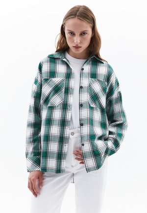Camisa de cuadros verde y blanca de manga larga, con cierre de botones, dos bolsillos frontales y un corte relajado, superpuesta sobre una camiseta blanca.