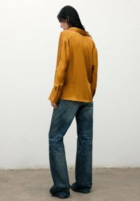 Camisa larga de satén color mostaza con mangas largas y cuello clásico, combinada con jeans acampanados de color azul desvanecido. Textura suave y ajuste entallado.