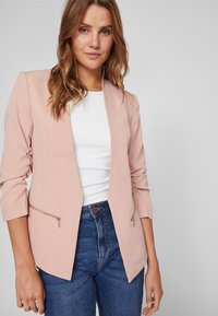 Blazer rosa empolvado con corte entallado, mangas largas y dos bolsillos frontales con cremallera, llevado sobre una parte superior blanca y jeans de denim.
