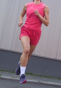 Top deportivo sin mangas rosa y pantalones cortos a juego con un patrón texturizado. Calcetines blancos y zapatillas oscuras con acentos rojos. Postura de carrera.