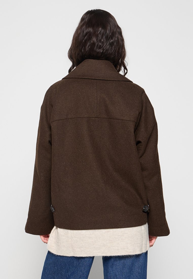 Giacca in misto lana marrone con colletto strutturato, dettaglio della spalla sul retro e due bottoni neri all'orlo, indossata sopra una camicia crema.