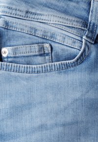 Tela de mezclilla azul claro con un bolsillo delantero visible y cierre de botón, que presenta un tejido texturizado y áreas ligeramente desgastadas.